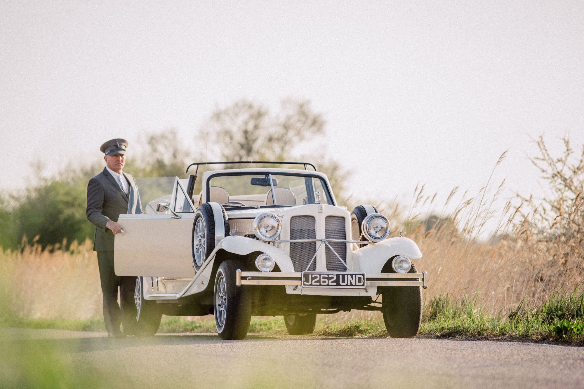 A cream vintage Beauford wedding car with chauffeur waiting beside an open door